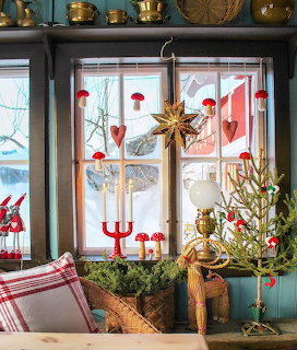 Window decorated with red heart ornaments, straw star, candles, and Christmas greenery with brass pots on shelf above