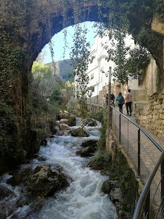 Stone arch over a rushing stream in Cazorla, Spain, with greenery and a metal walkway beside historic buildings