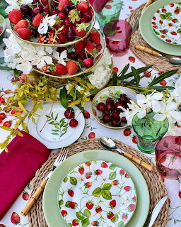 Top-down view of a table set with strawberry-patterned dishes, fresh berries, cheries, and white flowers on woven placemats