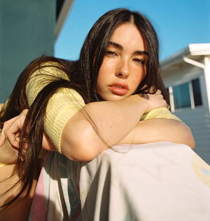 Close-up portrait of Madison Beer in a yellow top, leaning on her knees outdoors in sunlight
