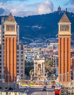 Barcelona cityscape showing two tall brick towers flanking a plaza with fountain and mountain backdrop