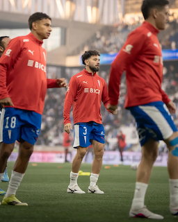 Three soccer players warming up on a stadium field in red tops and blue shorts.