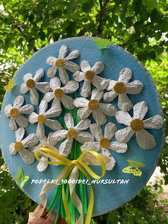 Handmade white paper daisy bouquet on a round blue board with yellow ribbon, held outdoors