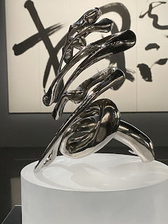 Shiny chrome steel sculpture with flowing curved forms on a white pedestal in front of caligraphy art