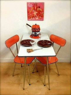 Retro kitchen table with orange chairs, brown fondue set, plates, and vintage poster on wall