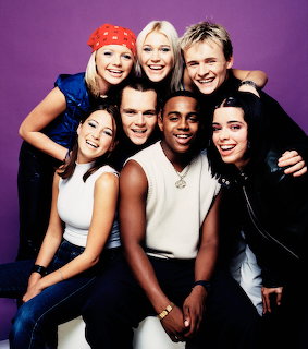 S Club 7 band members smiling and embracing in a studio portrait against purple background