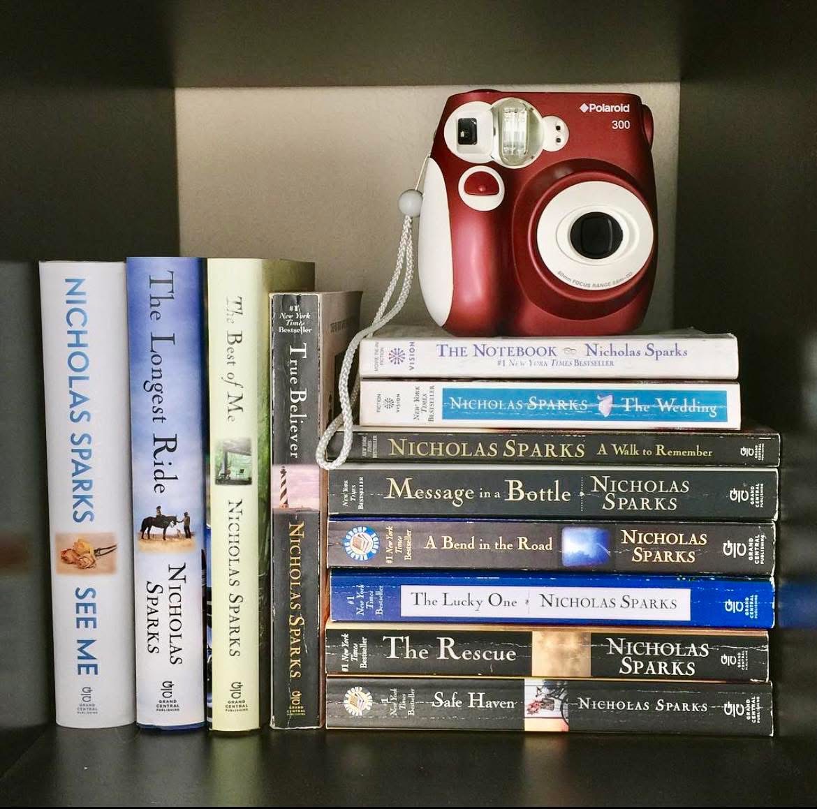 Nicholas Sparks books on a shelf with stacked novels and a red instant camera