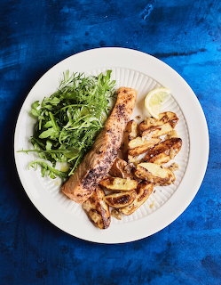 White plate with grilled salmon, roasted potatoes, arugula salad, and lemon on a blue background