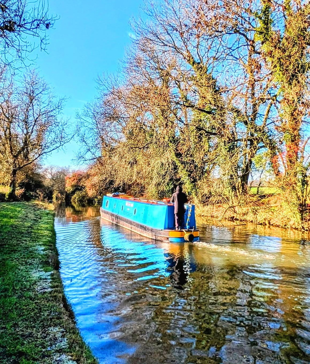 Blue narrowboat on a canal beside a grassy towpath and leafless trees