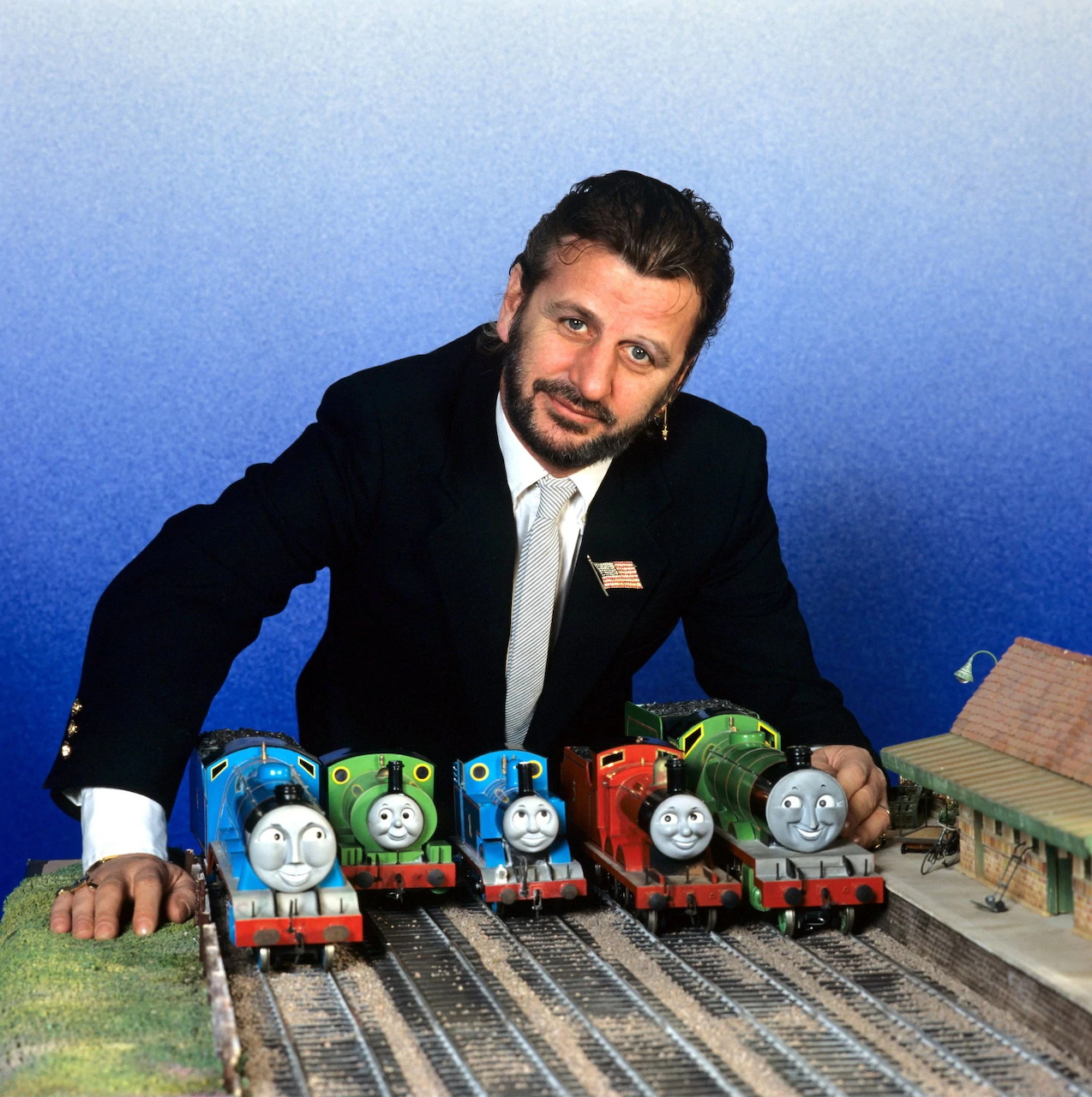 Ringo Starr behind toy train engines on model railway tracks with a blue backdrop