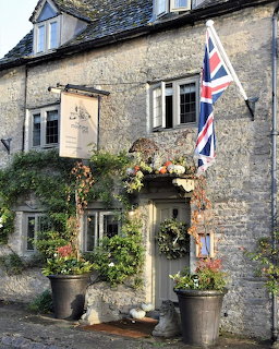 Stone cottage facade with Union Jack flag, hanging flower baskets, and poted plants by the door