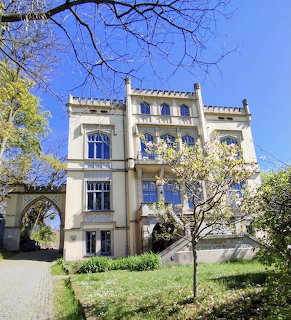 White three-story villa with decorative facade, gardens and bare trees against blue sky