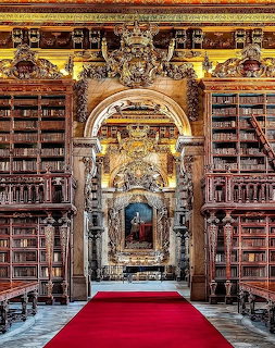 Baroque library hall with gilded shelves, dark wood, red carpet, and arched doorway with a portrait