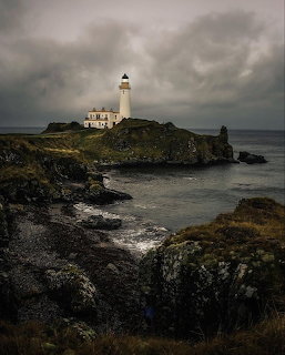 White lighthouse on a rocky green cliff above a dark sea under stormy grey clouds