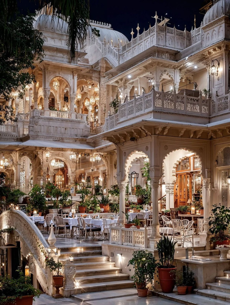 Illuminated ornate white Indian palace courtyard with carved balconies, dining area, and tropical plants at night