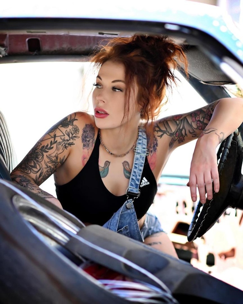 Tattooed redhead woman in denim overalls sitting inside a vintage car