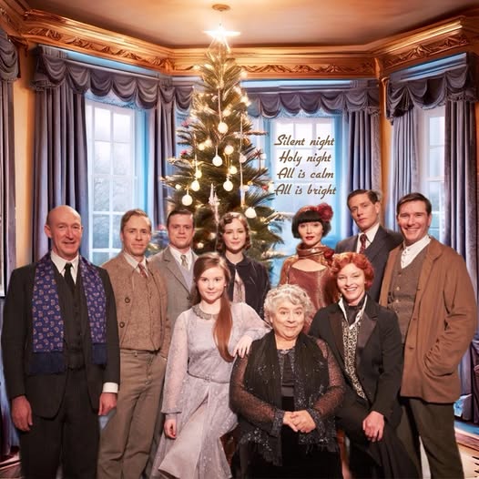 Nine people in period clothing pose indoors around a decorated Christmas tree