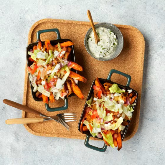 Two small black trays of loaded sweet potato fries with salad and dipping sauce on a cork board