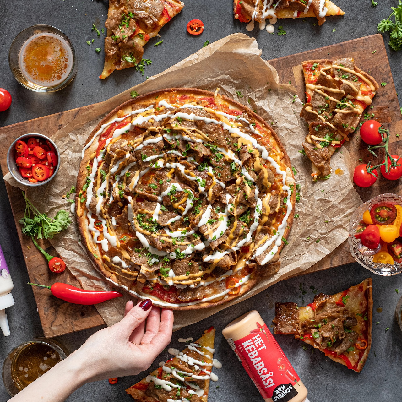 Overhead flatlay of a kebab pizza with spiral sauce, surrounded by tomatoes, chilies, and pizza slices on a dark table