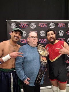 Man with glasses holding a championship belt posing with two wrestlers at an event
