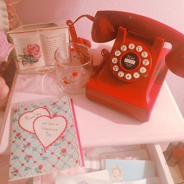Vintage red rotary telephone on a white table with a floral card, glass cup, and decorative boxes
