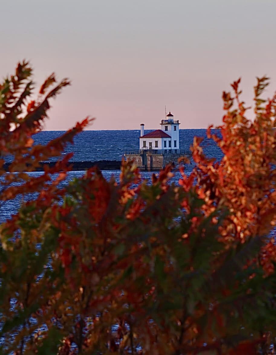 White lighthouse on horizon framed by red autumn foliage with blue water and pink sky