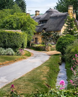 Stone cottage with thatched roof surrounded by green gardens and flowering plants along a curved path