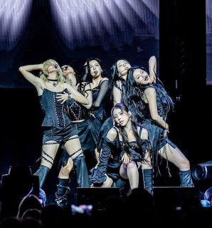 Five women in black performance outfits posing on a concert stage with blue lighting