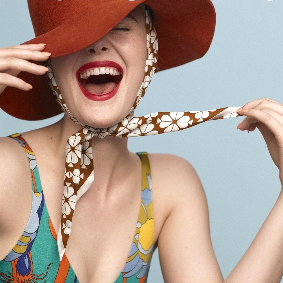 Woman wearing red sun hat and floral swimsuit laughing with head tilted back against light blue background