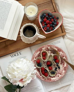 Overhead breakfast flat lay with berries in heart dish, coffee, open book, candle and white flower on wooden tray