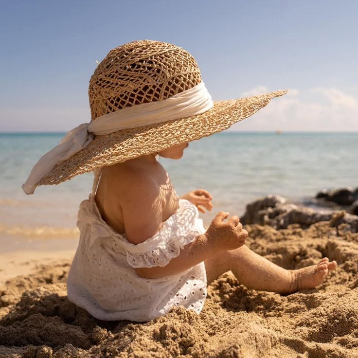 Toddler wearing a straw hat sitting in beach sand by the ocean