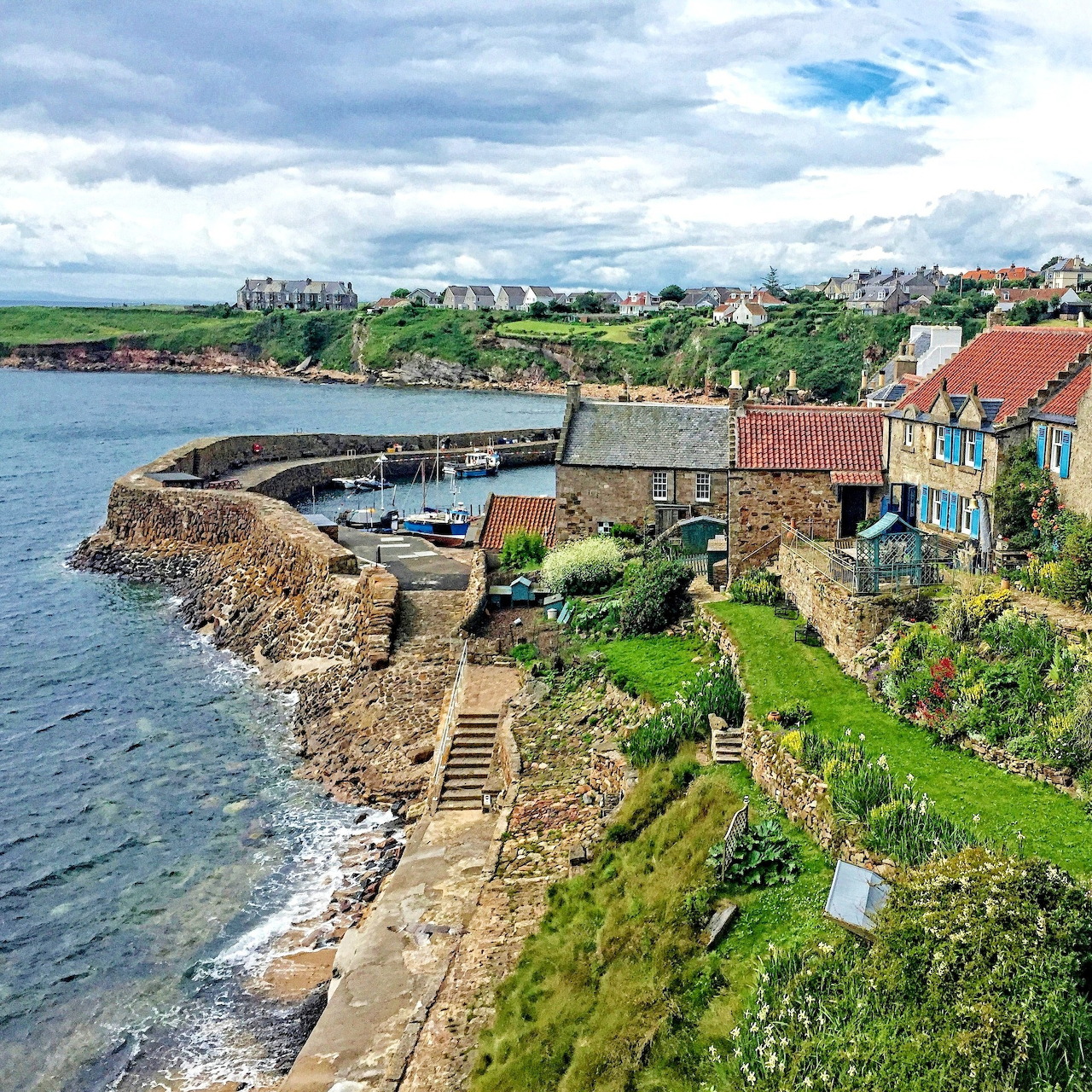 Aerial view of Crail fishing village harbor with stone walls, boats, colorful houses and cloudy sky