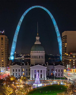 Blue-lit Gateway Arch frames illuminated courthouse dome at night with city buildings and streetlights