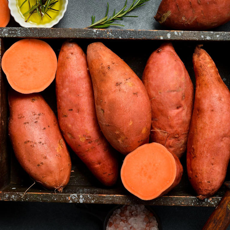 Whole and halved sweet potatoes with orange flesh arranged in a rustic wooden crate