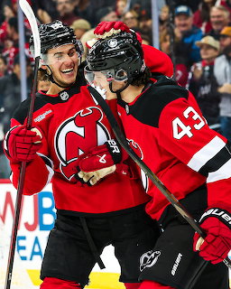 Two New Jersey Devils NHL players smiling and celebrating together on the ice in red jerseys