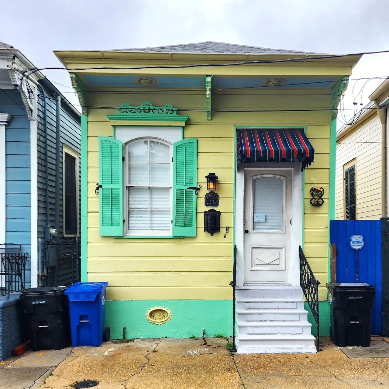 Small yellow house with turquoise shutters, white door, striped awning, front steps between blue and cream neighboring houses