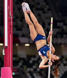Female pole vaulter inverted in mid-air during competition, gripping pole with arena seating in background