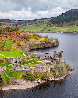 Aerial view of Urquhart Castle ruins on Loch Ness shoreline with green Scottish highlands and cloudy sky
