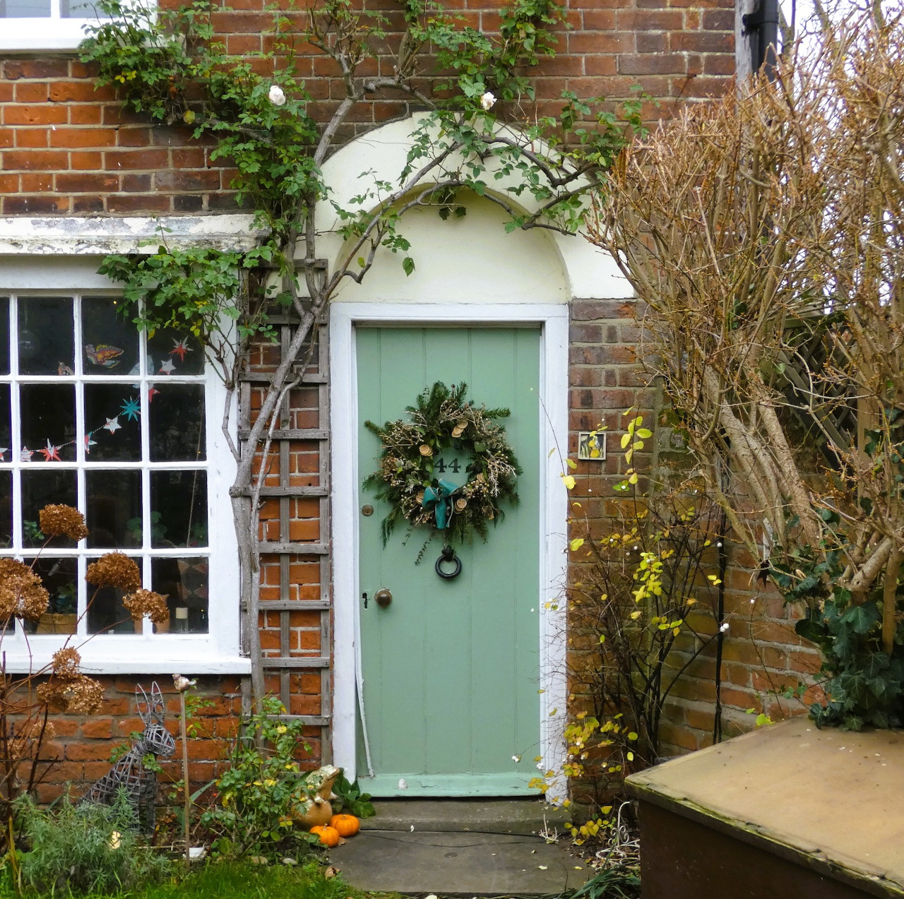 Mint green cottage door with white arched frame and wreath, surrounded by brick walls and garden plants