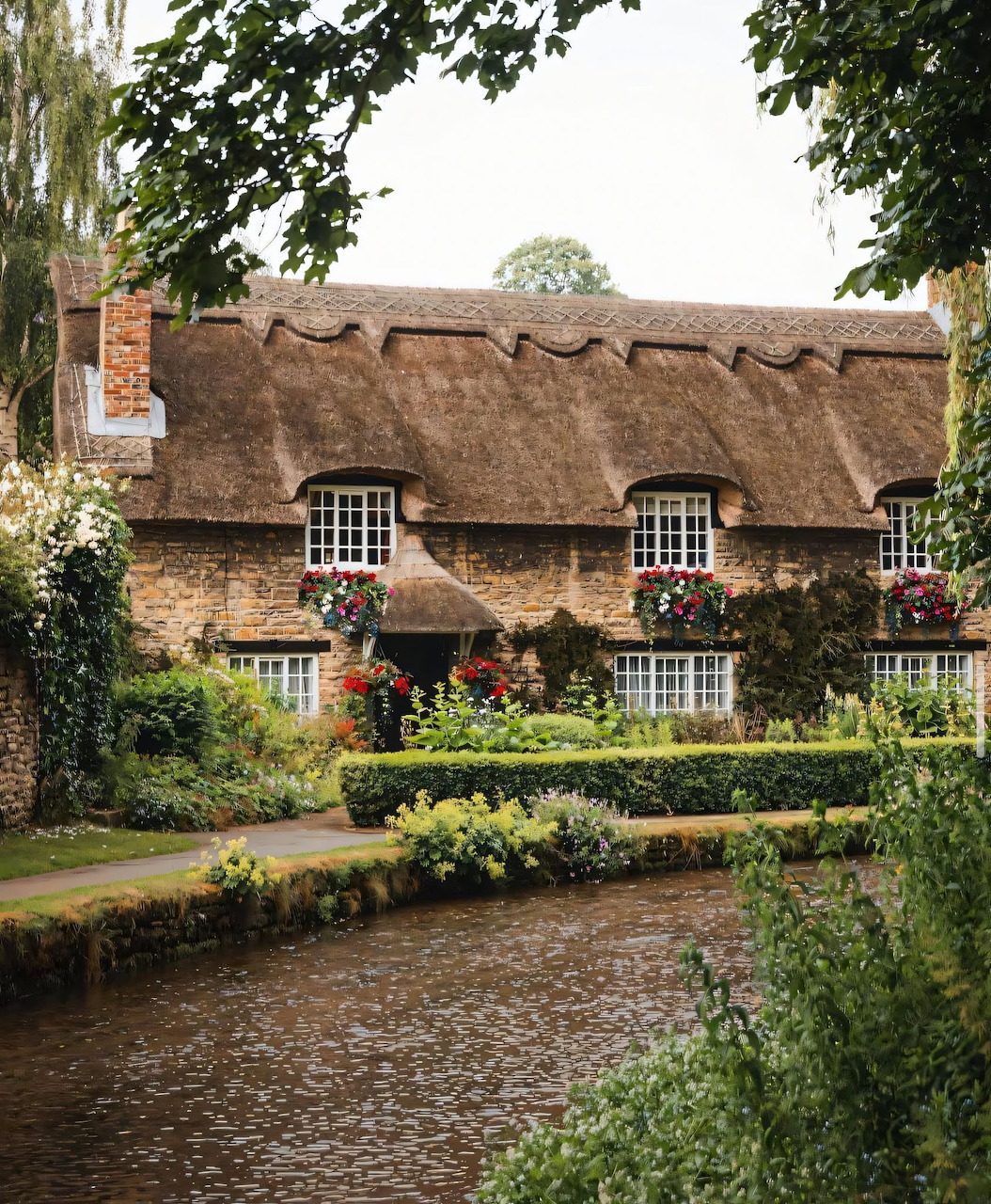 Thatched brick cottage with flowers and hedges beside a narrow waterway