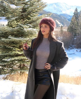 Woman in winter outfit posing in snow near an evergreen tree with mountains in the background