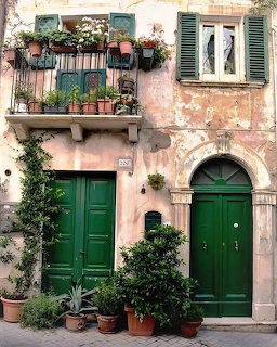 Weathered Italian building with two green doors, balcony with potted plants, and lush greenery
