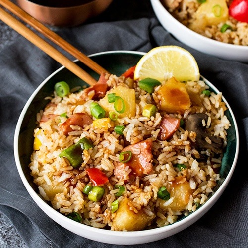 Bowl of Hawaiian fried rice with ham, pineapple, peppers, and lemon garnish, chopsticks resting on rim