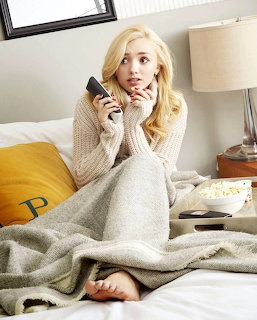 Blonde woman in beige sweater holding phone while sitting on bed in bright bedroom