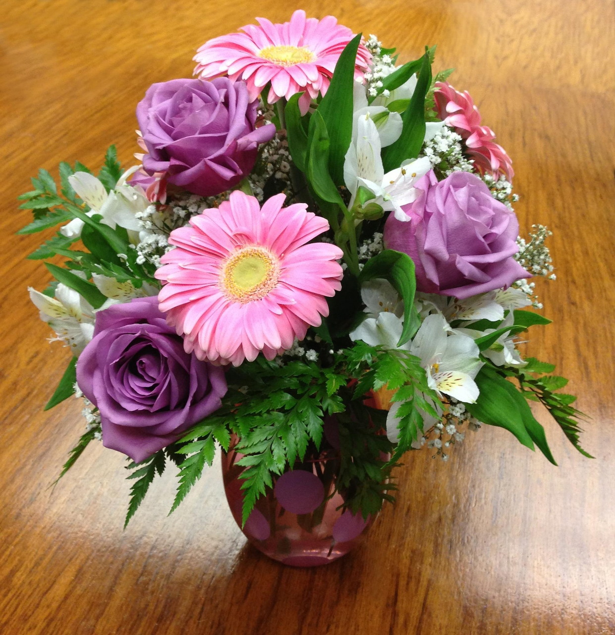 Bouquet of pink daisies, purple roses, and white flowers in a glass vase on a wooden table