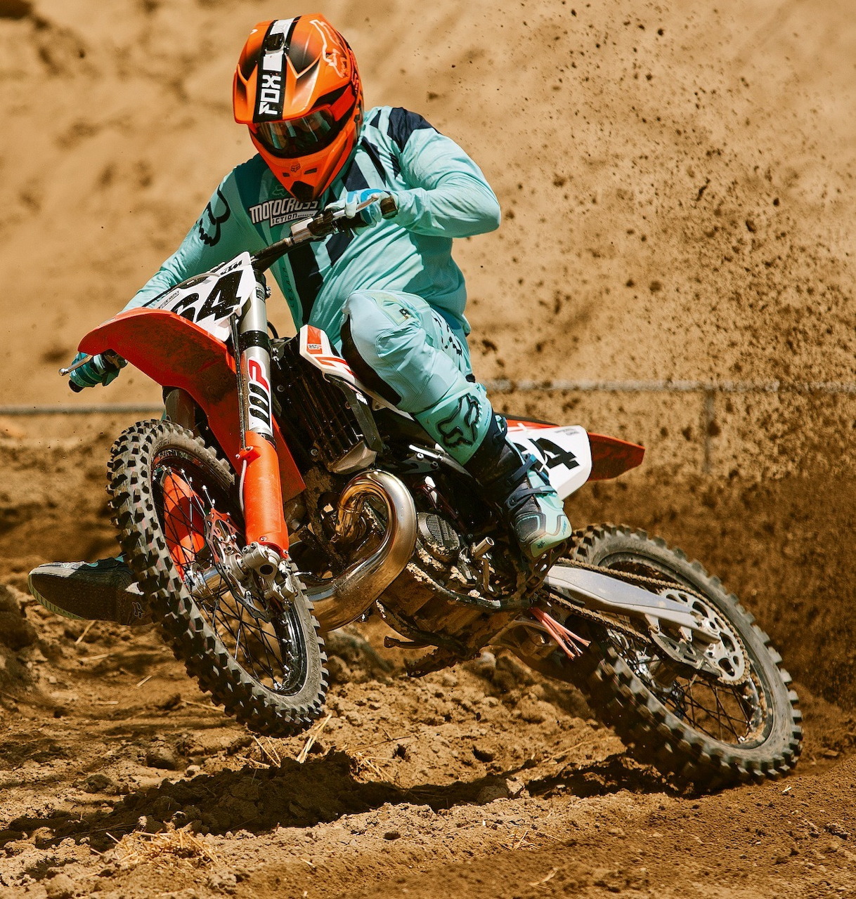 Motocross rider in teal racing gear riding orange dirt bike on sandy track
