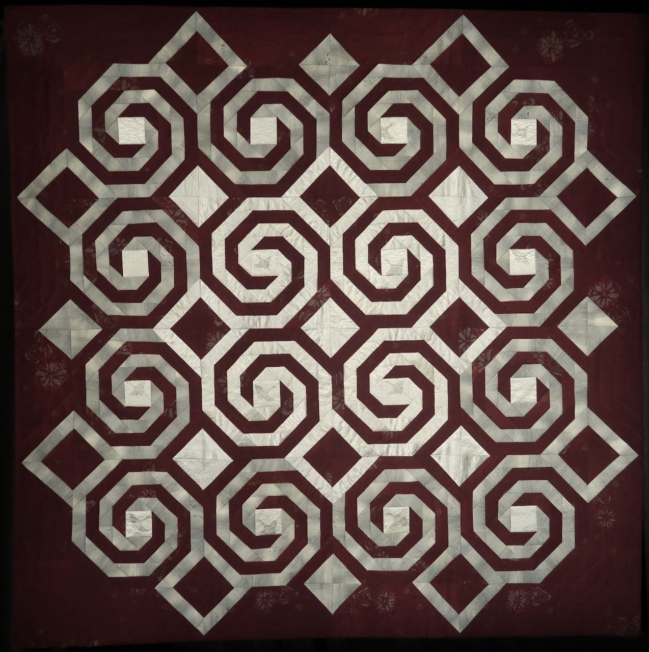 Symmetrical geometric pattern with repeating spiral and diamond shapes in burgundy, cream and gray