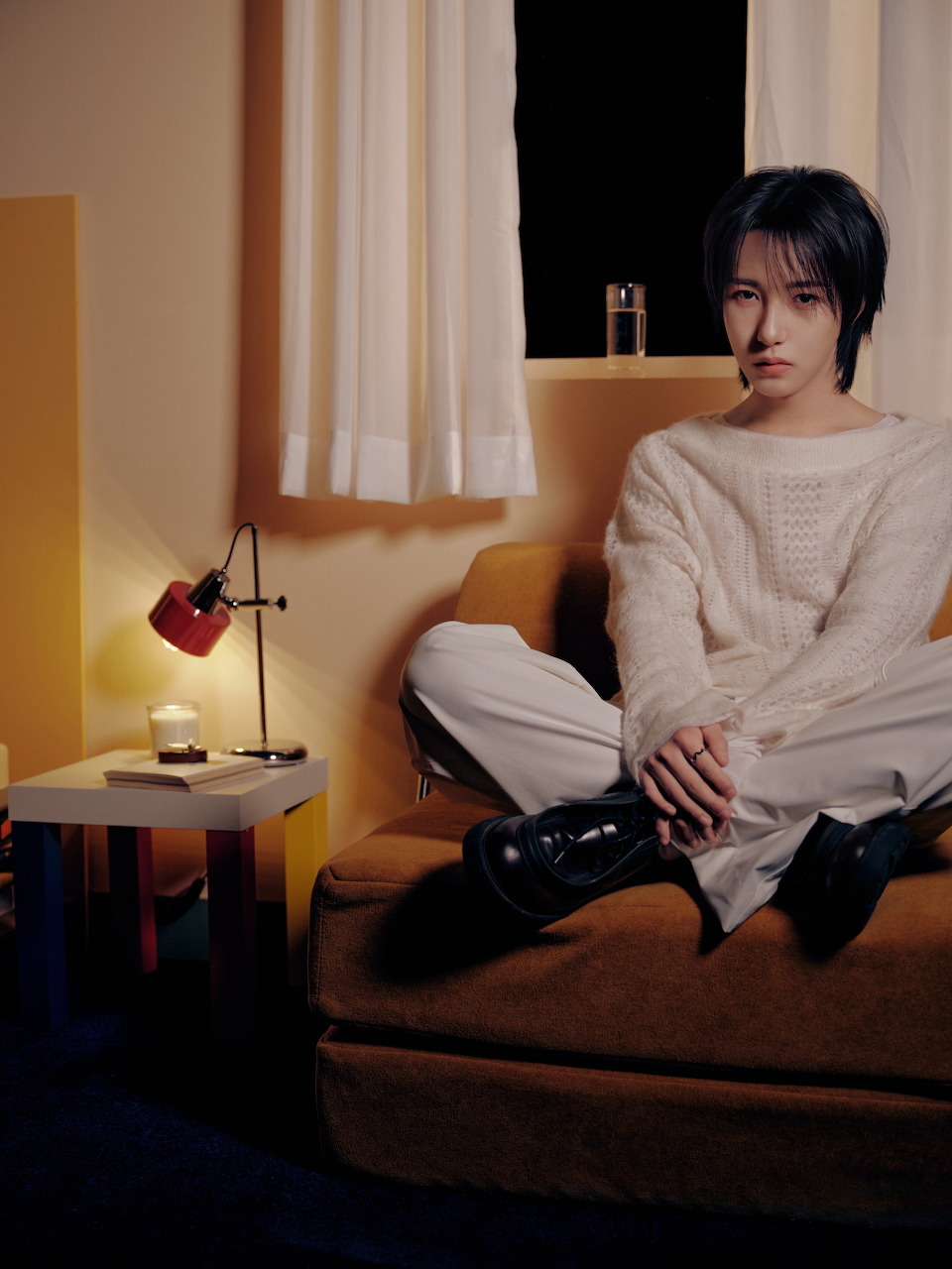 Young man in white sweater and black boots seated on an orange sofa in a cozy room with warm lighting