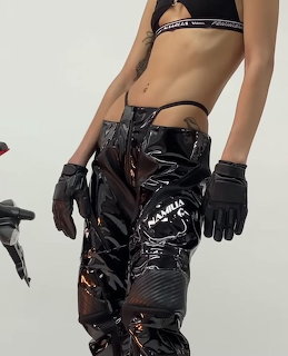 Woman in black shiny vinyl pants and racing gloves posing against a light background