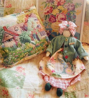Handmade rag doll in green cardigan next to cross-stitch cottage pilow on floral quilt